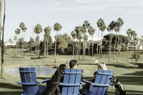 Three people sit on blue chairs by a golf course, with golf bags nearby. Palm trees and a pond are in the background.