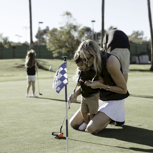 A woman and child on a golf course, focusing on putting near a hole with a checkered flag. Another person is in the background on the green.