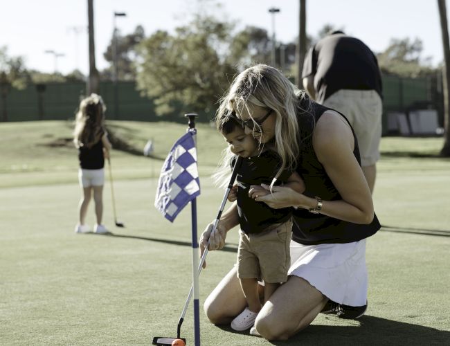 A woman and child on a golf course, focusing on putting near a hole with a checkered flag. Another person is in the background on the green.