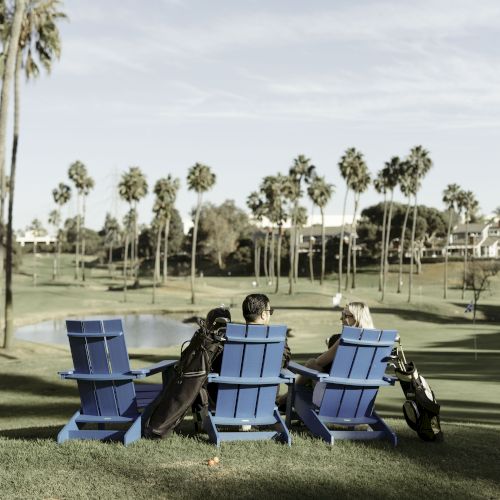 Three people sit on blue chairs with golf bags nearby, overlooking a scenic golf course with palm trees and a water feature in the background.