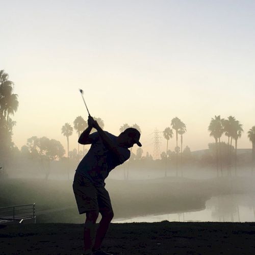 A silhouetted person is swinging a golf club on a misty golf course with palm trees and a body of water in the background.