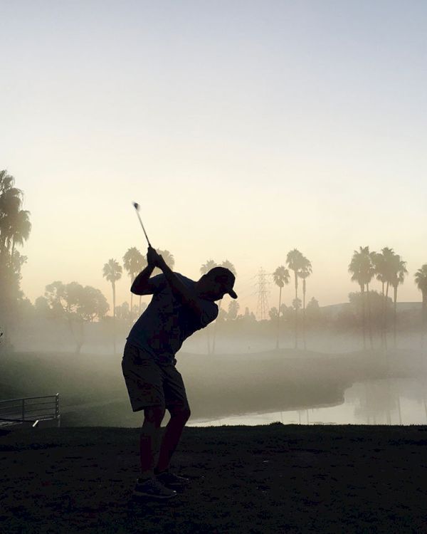 A silhouetted person is swinging a golf club on a misty golf course with palm trees and a body of water in the background.