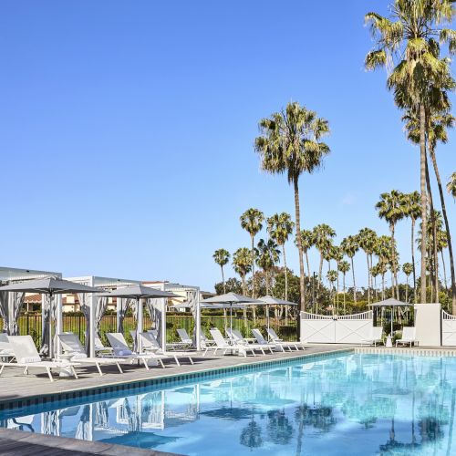 The image shows a swimming pool with lounge chairs and cabanas under a clear blue sky, surrounded by tall palm trees, creating a serene outdoor setting.