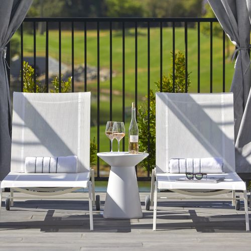 Two white lounge chairs with towels, a small table with wine glasses and a bottle, and outdoor curtains overlooking a green field.