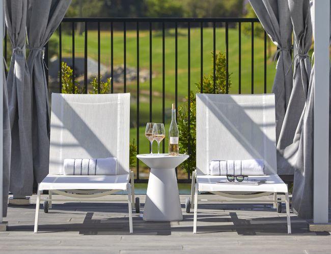 Two white lounge chairs with towels, a small table with wine glasses and a bottle, and outdoor curtains overlooking a green field.