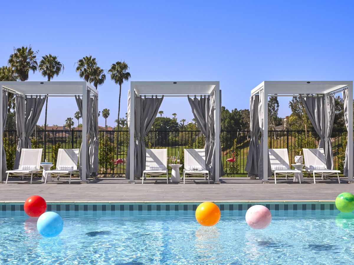 The image shows a swimming pool with colorful beach balls and three cabanas with lounge chairs under a clear blue sky, ending the sentence.