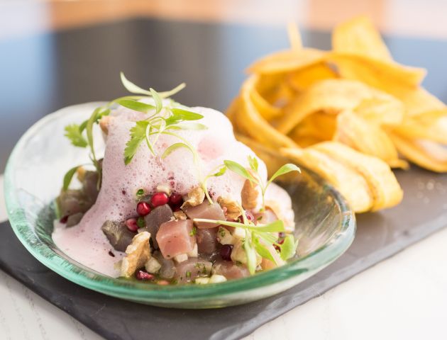 A plate of diced seafood topped with a pink foam, garnished with greens, and accompanied by a side of plantain chips.