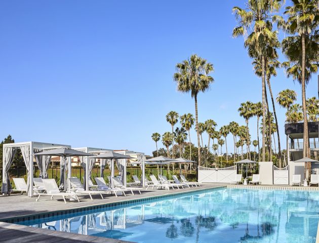 A serene outdoor pool with lounge chairs and white canopy beds, surrounded by palm trees under a clear blue sky.