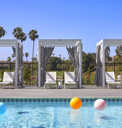 A pool with colorful beach balls, behind which are three cabanas flanked by palm trees under a clear blue sky.