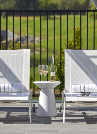 Two lounge chairs sitting under a canopy with a small table between them, holding wine glasses and a bottle, overlooking a green landscape.