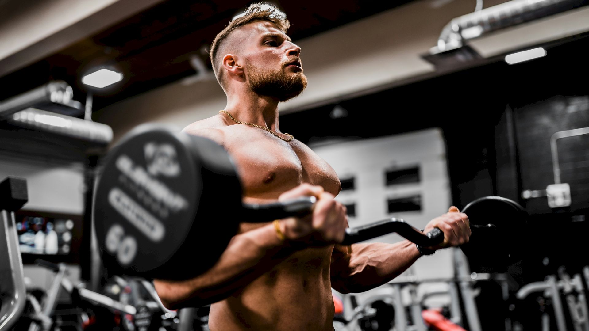 A person is lifting a barbell in a gym environment with various gym equipment in the background.
