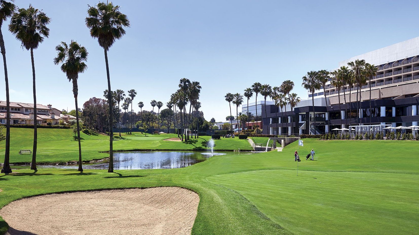 The image shows a golf course with palm trees, a sand bunker, a pond, and buildings in the background. Two golfers can be seen playing.