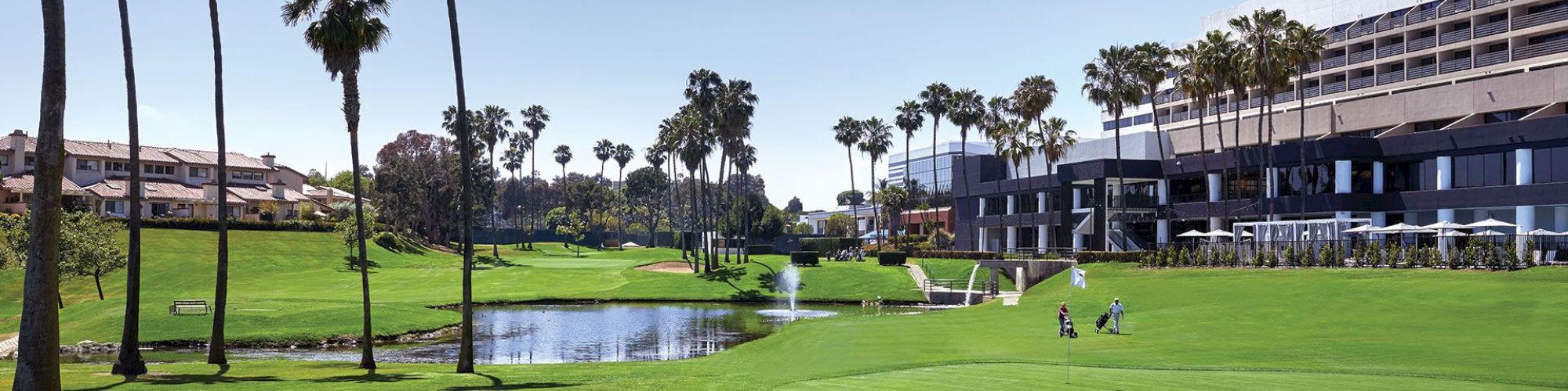 The image shows a golf course with palm trees, a sand bunker, a pond, and buildings in the background. Two golfers can be seen playing.