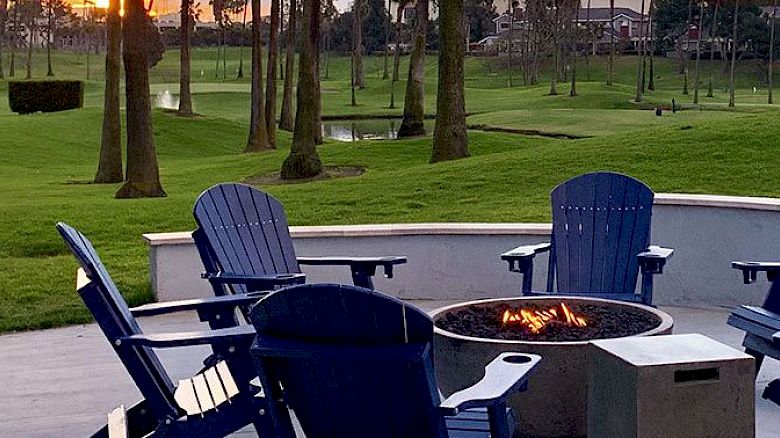 The image shows a patio with blue Adirondack chairs around a fire pit, overlooking a golf course at sunset.