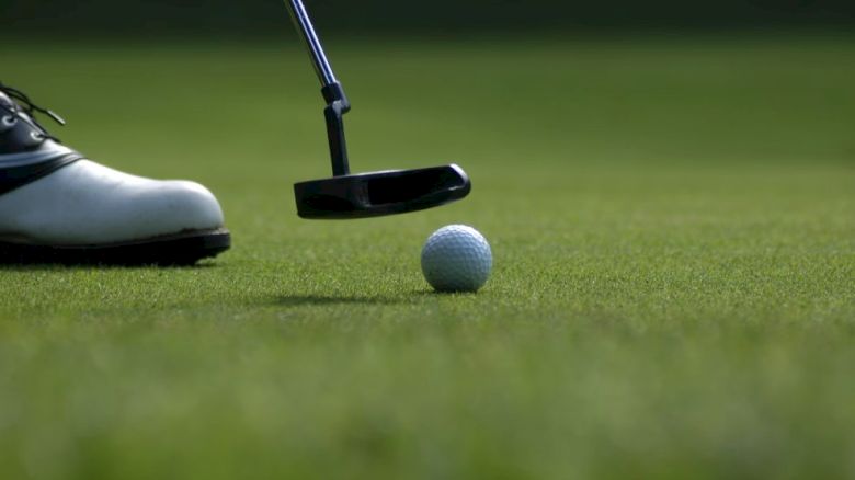 The image shows a close-up of a person putting a golf ball on a green field, with a golf club and part of a shoe visible.