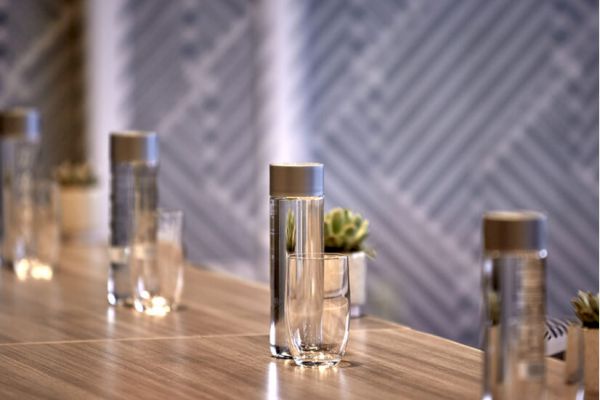 The image shows a wooden table with sleek glass bottles and glasses placed on it, with a patterned background and small decorative plants.