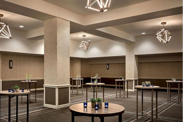 The image shows a modern, spacious conference room with round high tables, geometric ceiling lights, and minimalist decor, including small plants on tables.