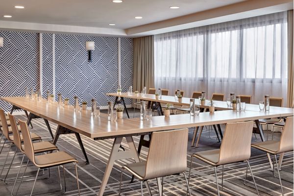 A modern conference room with a long wooden table, chairs, water bottles, and glasses, featuring a geometric patterned wall and large windows.