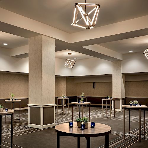 An empty event space with modern geometric lighting, round tables with small plants and candles, and a patterned carpeted floor.