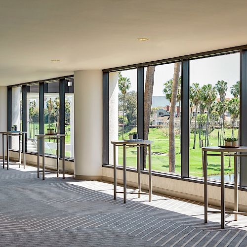 A brightly lit room with tables along large windows overlooks a green landscape with palm trees and a lake in the distance.