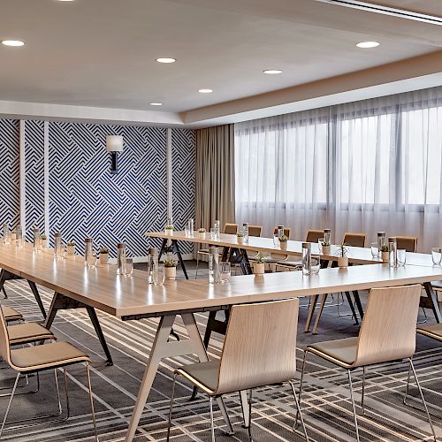 A modern conference room with a U-shaped wooden table, numerous chairs, water bottles, large windows with curtains, and patterned walls.