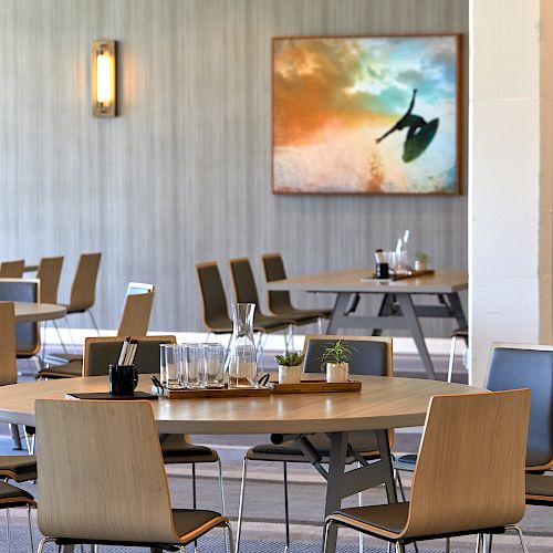 The image shows a modern dining area with neatly arranged wooden tables and chairs, glassware, and a wall painting in the background.