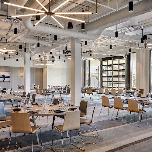 A modern dining area with multiple tables and chairs, set with plates and glasses. Ceiling features unique lighting fixtures, and large windows.