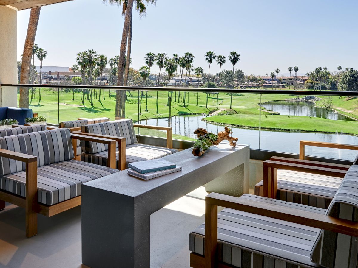 A patio area with striped cushioned chairs, a table, and a view of a golf course with palm trees and a pond in the background.