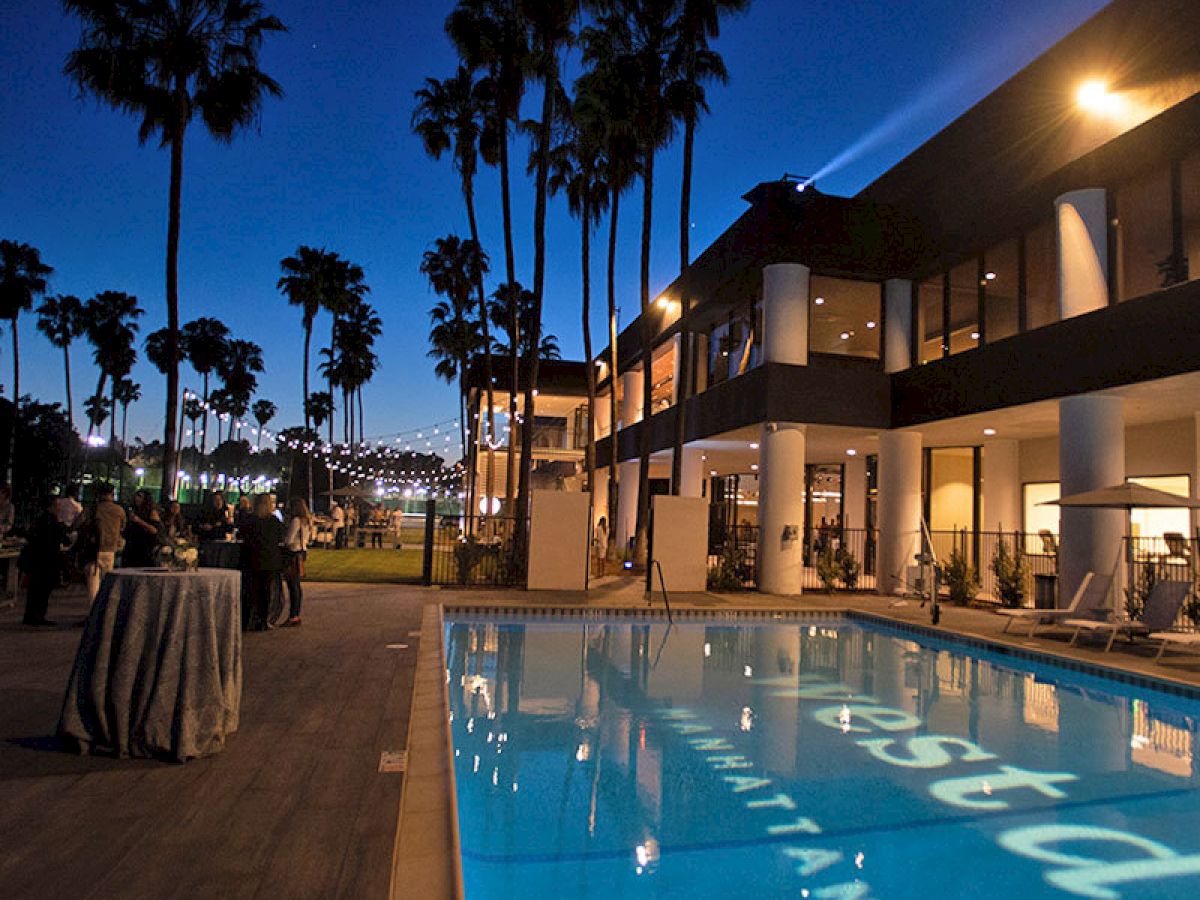 The image shows a luxurious outdoor pool area at night with palm trees, people socializing, and a modern building in the background.