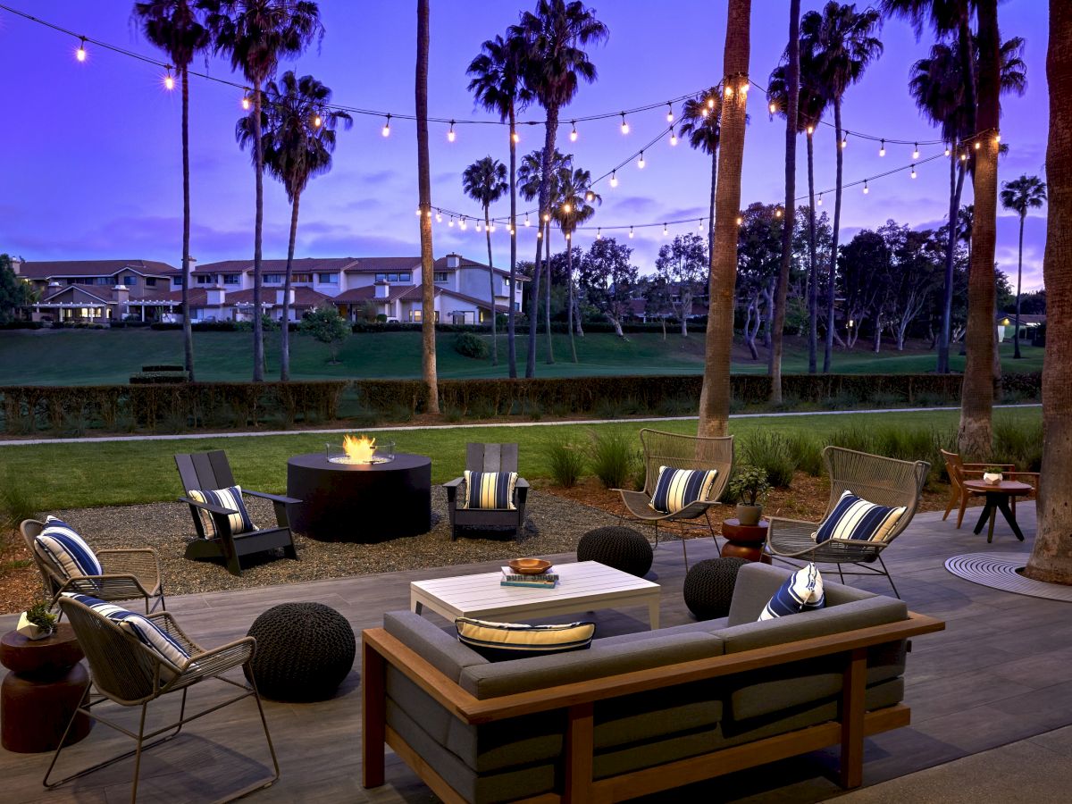 A stylish outdoor seating area is illuminated with string lights near tall palm trees, featuring a fire pit and modern furniture at dusk.