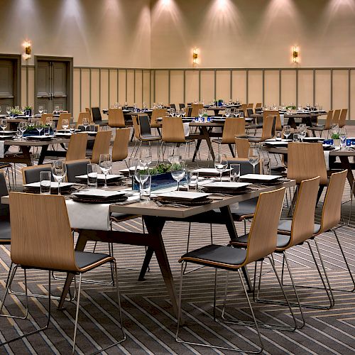 A banquet room is set up with round tables and wooden chairs, each table arranged with plates, glasses, and napkins ready for an event.