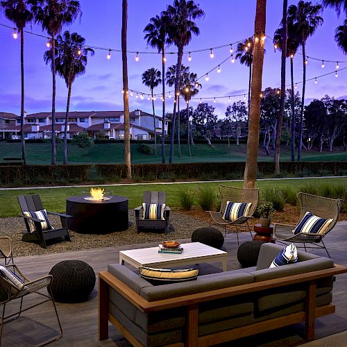 Outdoor seating area with cushioned chairs, a couch, and a fire pit under string lights. Palm trees and buildings can be seen in the background.