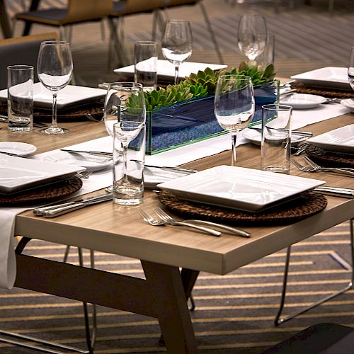 A dining table set for six with plates, wine glasses, cutlery, and a centerpiece featuring green plants in a rectangular container, in a modern setting.