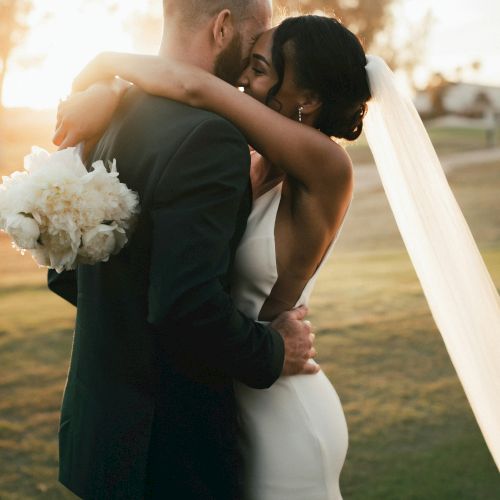 A couple shares an intimate embrace and kiss during their wedding ceremony at sunset. The bride holds a bouquet and wears a veil.