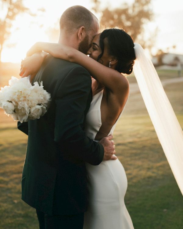 A couple shares an intimate embrace and kiss during their wedding ceremony at sunset. The bride holds a bouquet and wears a veil.