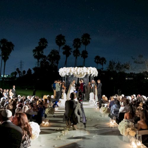 The image shows an outdoor nighttime wedding ceremony with guests seated and a couple under a floral arch, surrounded by palm trees.