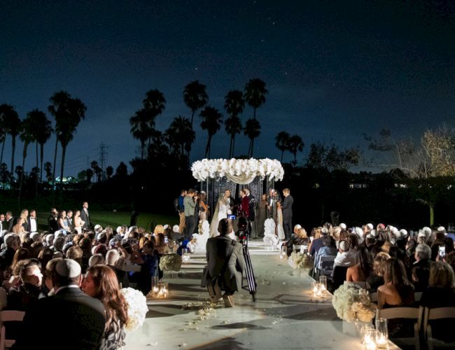 The image shows an outdoor nighttime wedding ceremony with guests seated and a couple under a floral arch, surrounded by palm trees.