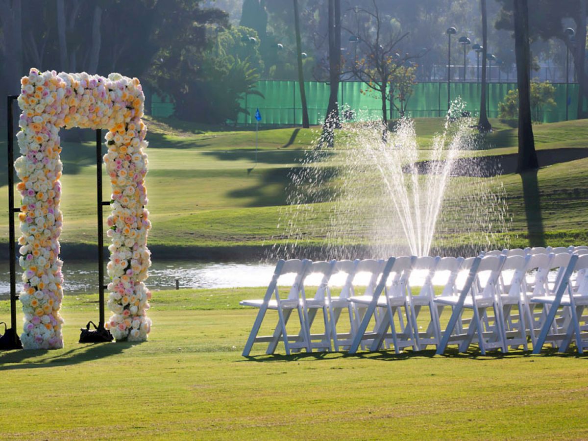 An outdoor wedding setup featuring a decorative floral arch, a row of white chairs, and a fountain in the background on a well-maintained lawn.