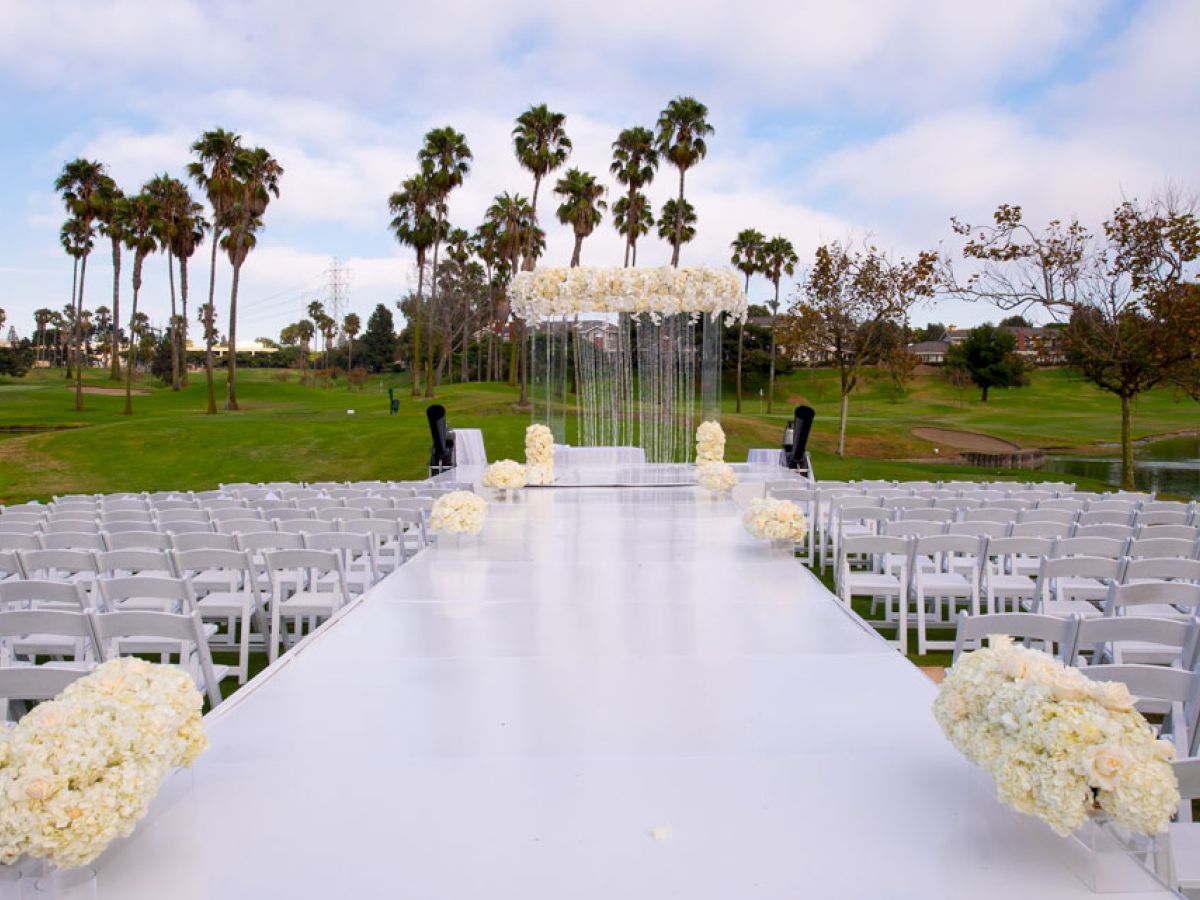 The image shows an outdoor wedding setup with white chairs arranged on either side of a white aisle, decorated with white flowers, likely on a golf course.