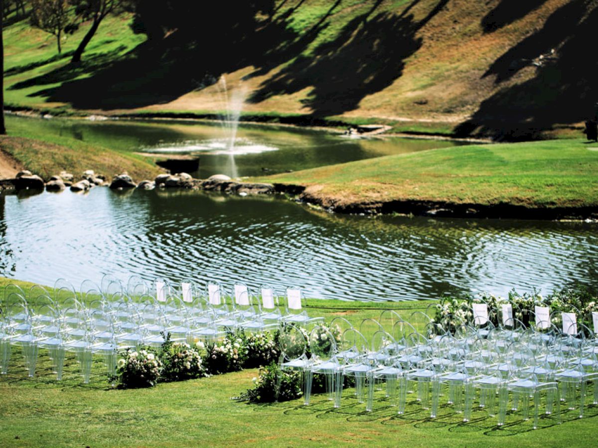 The image shows an outdoor wedding setup by a serene pond with a fountain, featuring rows of transparent chairs arranged on the grass.