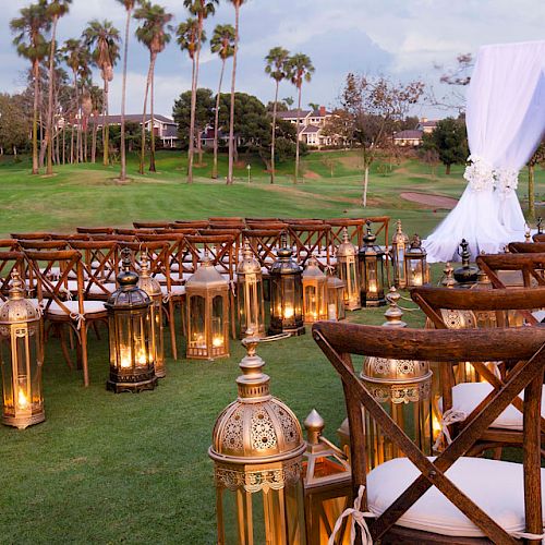 An outdoor wedding setup with wooden chairs, lanterns, and a white draped arch on a lawn with tall palm trees and a building in the background.