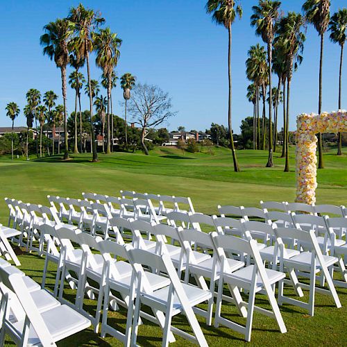 White chairs are set up in rows on a green lawn with palm trees and an arch decorated with flowers, indicating a wedding ceremony location.