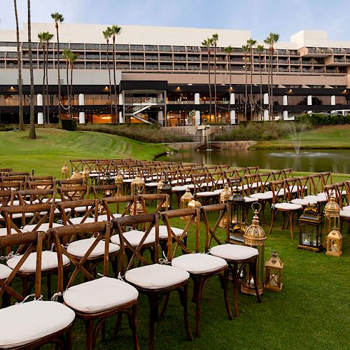 An outdoor wedding setup with rows of wooden chairs and lanterns on a lawn, facing a building and small lake in the background.