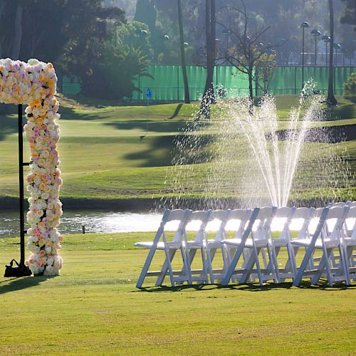 An outdoor wedding setup featuring a floral arch and rows of white chairs next to a fountain on a well-manicured lawn.