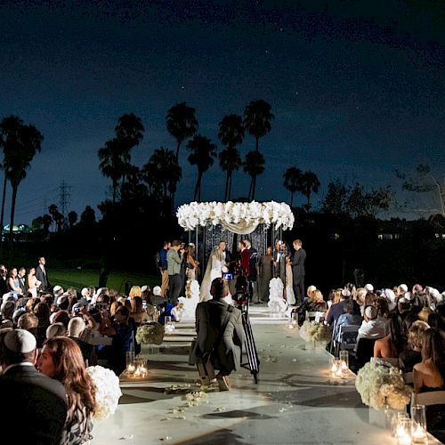 A nighttime outdoor wedding ceremony with guests seated and a couple standing under a floral canopy, surrounded by palm trees and string lights.