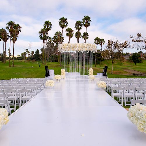 The image shows an outdoor wedding setup with white seating arranged on either side of an aisle with floral decorations under a floral arch with a backdrop.