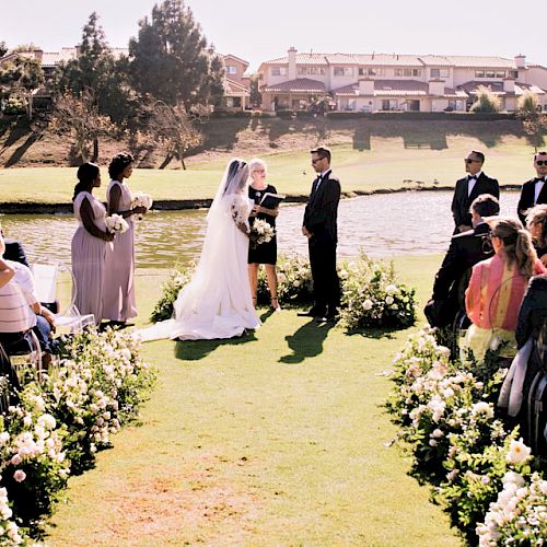 A wedding ceremony outdoors with a bride and groom at the altar, surrounded by guests, bridesmaids, and groomsmen near a lake under sunny skies.