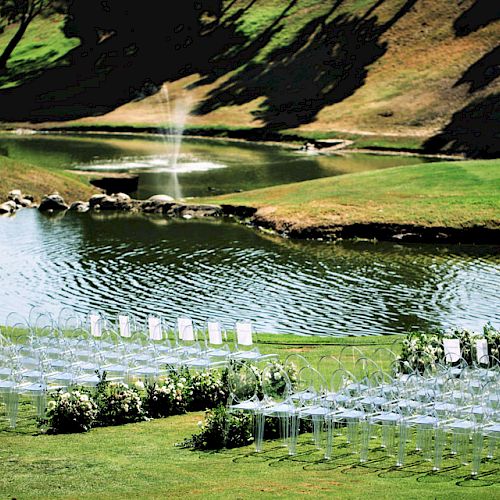 Rows of transparent chairs arranged outdoors in front of a pond, possibly for an event, with a fountain in the background and greenery around.