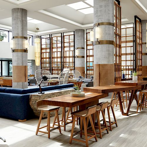 A modern, spacious lounge area with wooden tables, chairs, a blue sofa, hanging rattan chair, and large lamps, featuring concrete pillars and ample lighting.