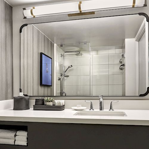 A modern bathroom vanity with a large mirror, double sink, towels, toiletries, and a view of a shower area reflected in the mirror.